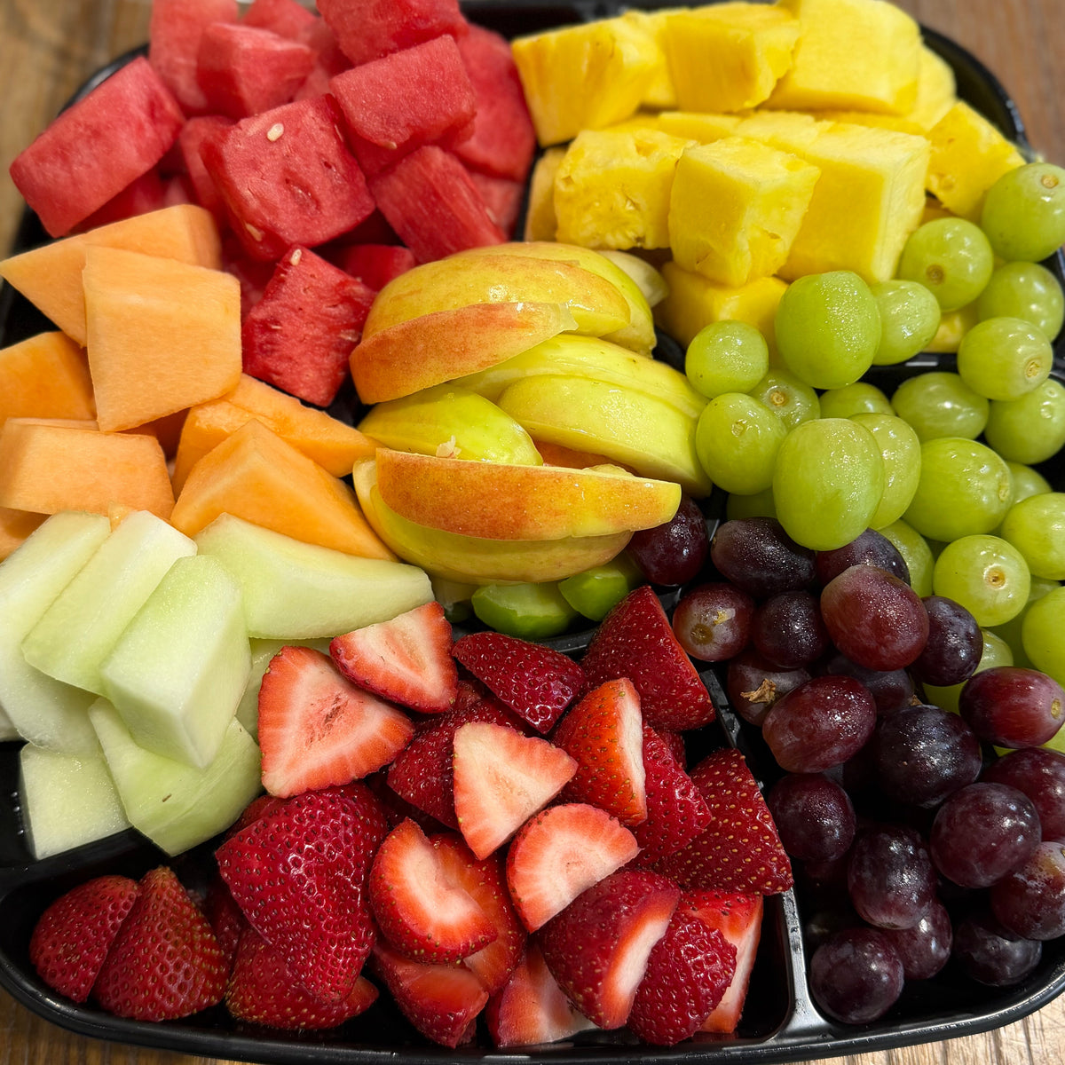 Assorted fruits including watermelon, pineapple, apples, grapes, and strawberries on a black tray.