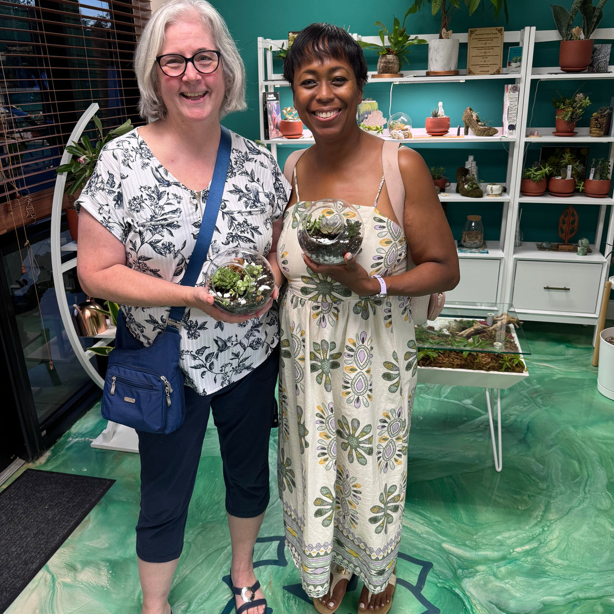 Two women holding small plants in a room with shelves of plants.