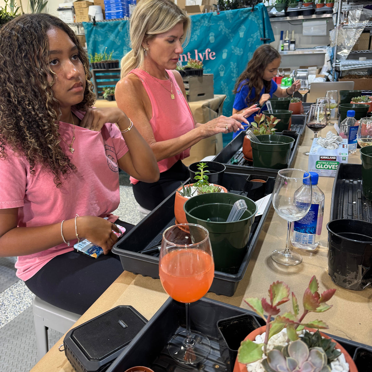 Two women in a workshop setting with plants and tools, one holding a glass of orange juice.