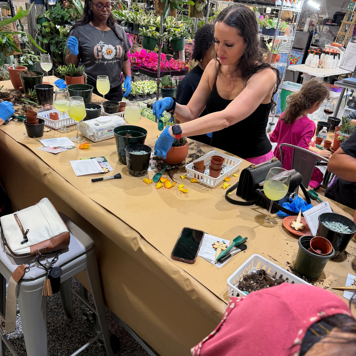 Woman potting plants at a table with other people in a garden center setting.