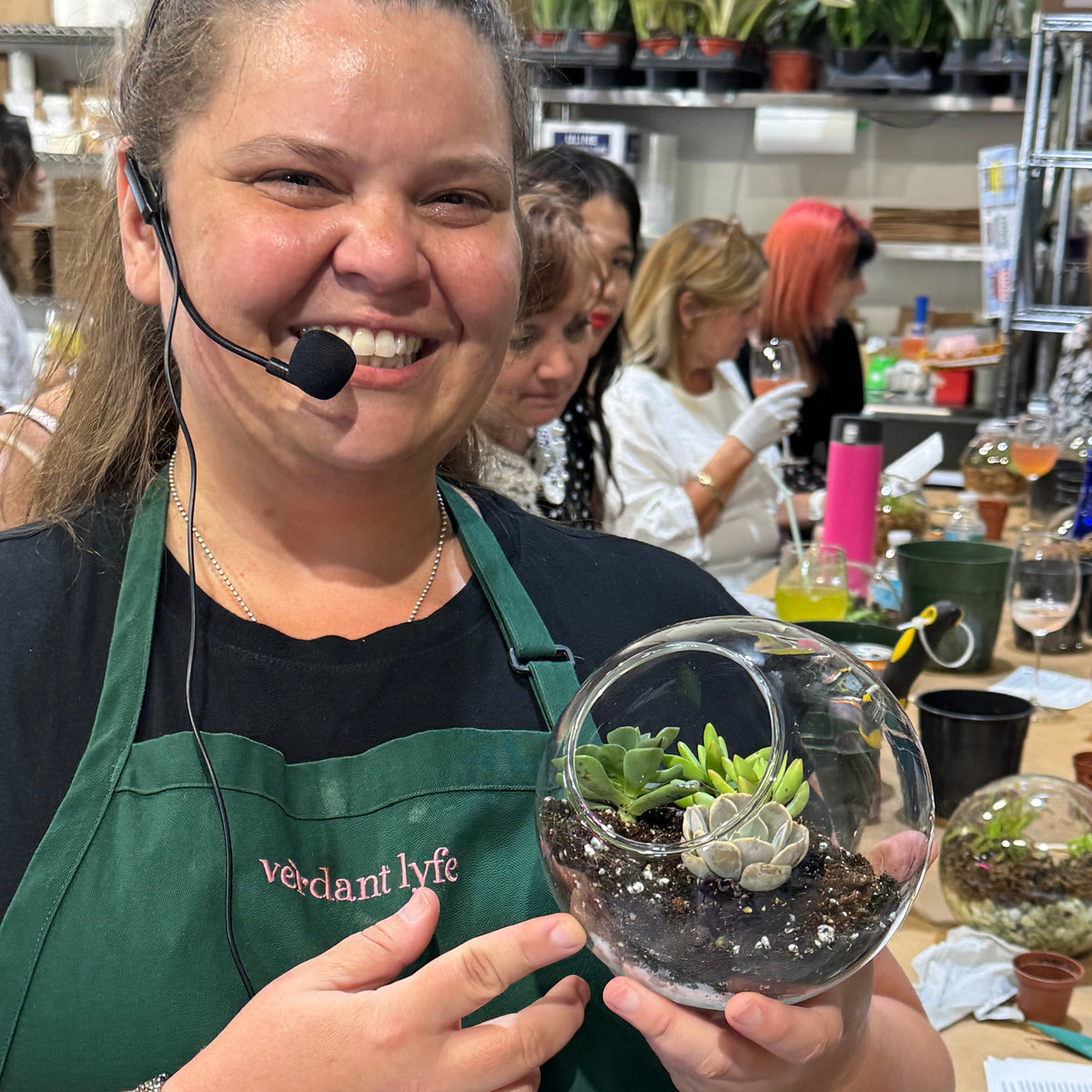Person in a green apron holding a terrarium with plants, surrounded by other people and materials in a workshop setting.